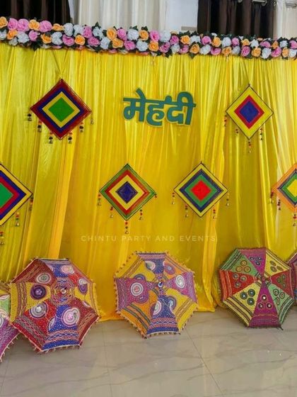 A colorful Rajasthani theme decoration for a Mehndi ceremony. The setup features a bright yellow backdrop, decorative umbrellas, and colorful hanging tassels.