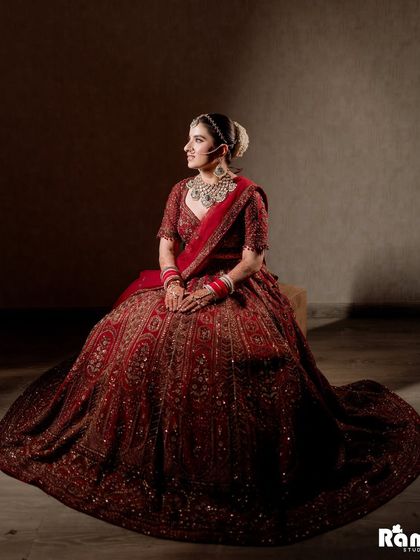 A regal portrait of the bride seated, her voluminous red lehenga spread around her. The dark background makes her bridal attire stand out beautifully.