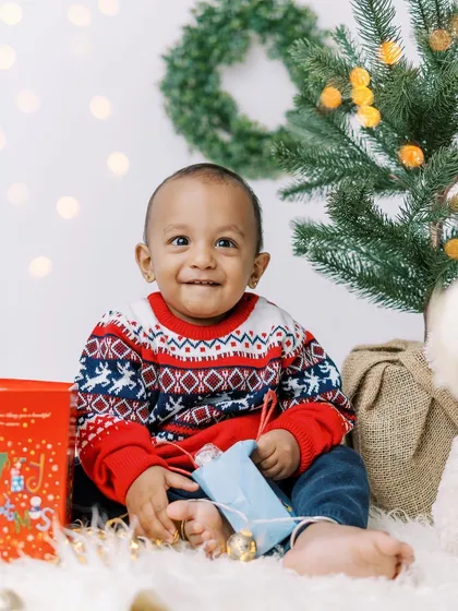 A baby boy in a festive sweater, smiling brightly next to the Christmas tree. His joy is infectious.