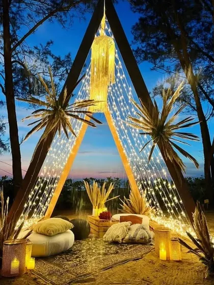 A stunning bohemian-style triangular cabana on a beach at sunset. The structure is draped in fairy lights, creating an incredibly romantic and picturesque setting.
