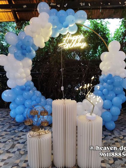 A simple and elegant birthday setup with a circular balloon arch in blue and white. The decor is accented by a neon "Happy Birthday" sign and minimalist white fluted plinths.