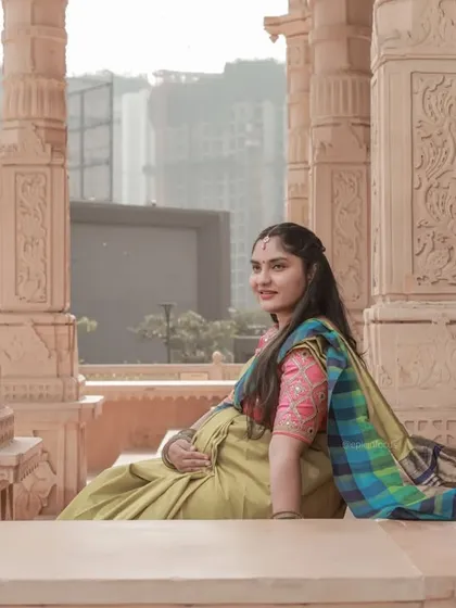 A serene solo portrait of a mom-to-be in a colorful saree, resting against the carved pillars of a beautiful outdoor location.