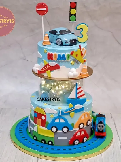 This transport-themed cake is a dream for any vehicle lover. It's a two-tier design with a light-up spacer, featuring a 3D car topper, fondant airplanes, boats, and a Thomas the Train figure circling the base.
