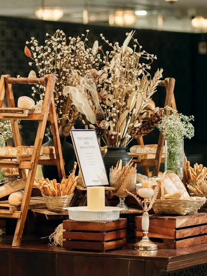 A rustic bread and cheese station, complete with a variety of artisanal loaves, breadsticks, and a warm cheese fondue. The wooden crates and dried flowers add to the charming, rustic aesthetic.