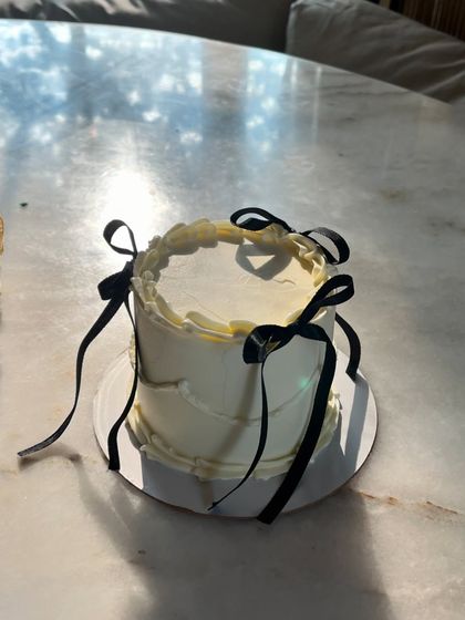 A minimalist coquette cake basking in the sunlight. The simple white-on-white piping and delicate black bows create a chic and sophisticated look.