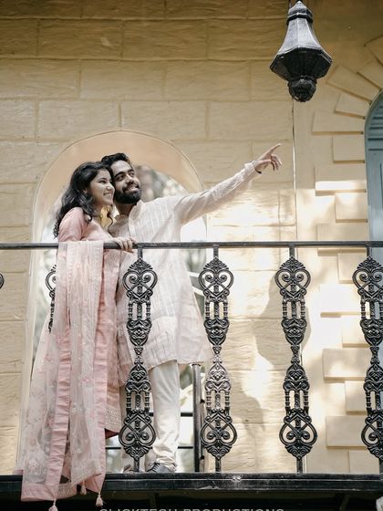A couple shares a romantic moment on a balcony with ornate railings, dressed in elegant, light-colored traditional Indian wear.