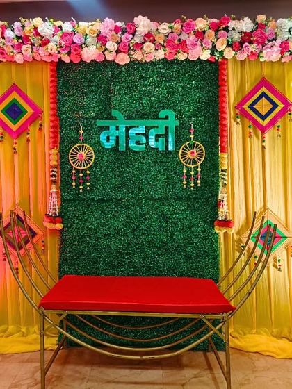 Here is a recent Mehndi decoration I did, featuring a green grass wall backdrop with a "Mehndi" sign in Hindi. The look is completed with colorful hangings and a red bench for the bride.