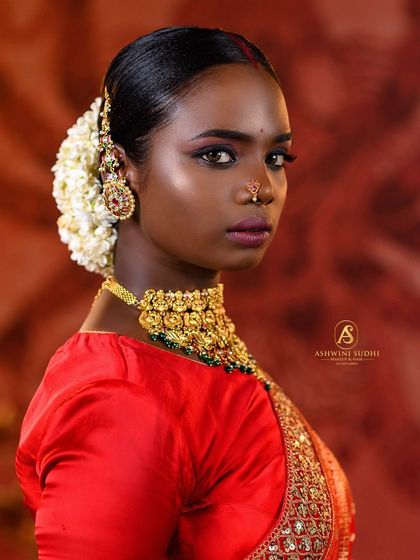 A striking profile shot of a model in a traditional bridal look. The focus is on the elegant hairstyle adorned with flowers and the classic makeup, captured with soft, flattering light.
