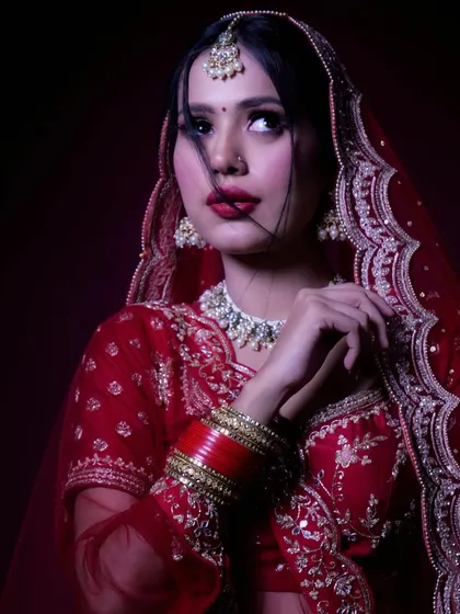 A dramatic, thoughtful pose of the bride looking away from the camera. The lighting sculpts her features and highlights the beautiful embroidery on her red bridal outfit.