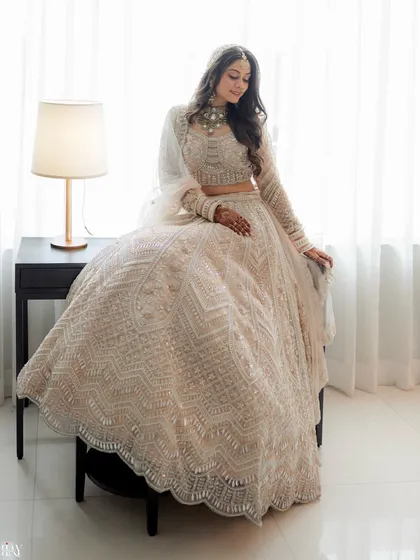 The bride looking serene and elegant in her white lehenga, seated in a beautifully lit room.