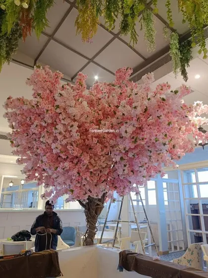 This stunning, large-scale artificial pink cherry blossom tree acts as a magnificent centerpiece in a cafe, offering a vibrant and unique ambiance that attracts customers.