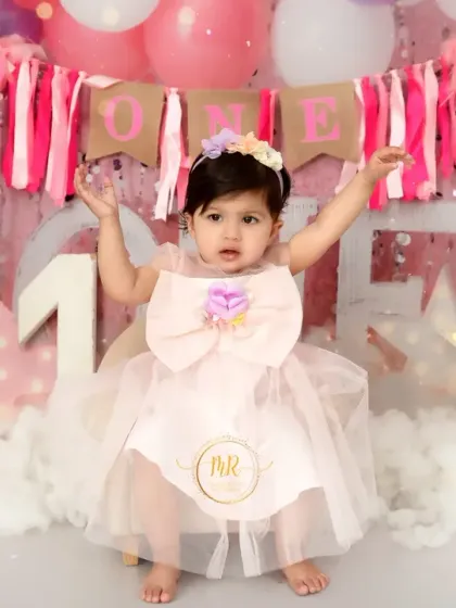 A one-year-old girl in a cute pink dress with a big bow, celebrating with arms up in a festive pink-themed birthday setup.