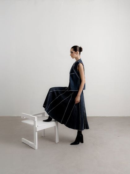 A profile shot from a lookbook for Nakateki, emphasizing the architectural lines of the denim vest and pleated skirt. The minimalist setting keeps the focus entirely on the garment's silhouette.