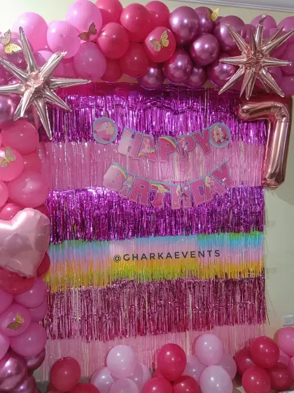 A vibrant pink Barbie-themed 7th birthday. The decor features a shimmering tinsel backdrop in pink and purple, a pink balloon arch, and a large foil number '7'.