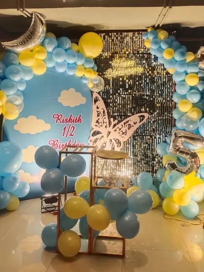 A sky-themed half-birthday decoration for a baby boy. The backdrop features a blue sky with clouds, framed by a yellow and blue balloon arch, a crescent moon foil balloon, and a number '5' foil balloon to mark the 5th-month celebration.
