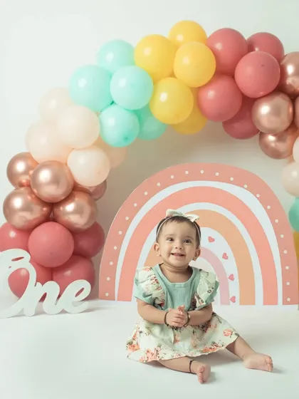 The birthday girl sits happily in front of a beautiful rainbow balloon arch, ready for her first birthday celebration.