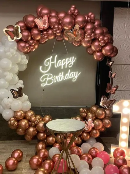 An elegant first birthday setup with a rose gold and white color scheme. The circular balloon arch is accented with butterfly details and surrounds a 'Happy Birthday' neon sign, with a marquee number '1' on the side.