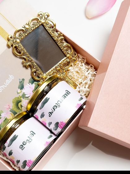 Introducing the 'Thamara' hamper in its elegant pink box. This gift focuses on simplicity and tradition, featuring jars of snacks and a vintage-style valkannadi (hand mirror).
