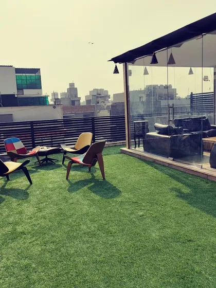 After: The same rooftop transformed with a lush artificial lawn and a modern glass-enclosed lounge, furnished with stylish, colorful chairs.