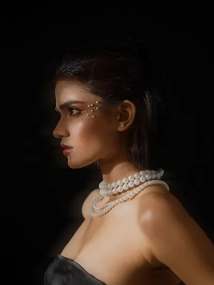 A dramatic, low-key beauty portrait in profile. The lighting highlights the contours of the model's face and the shimmer of the pearl necklace against the dark background.