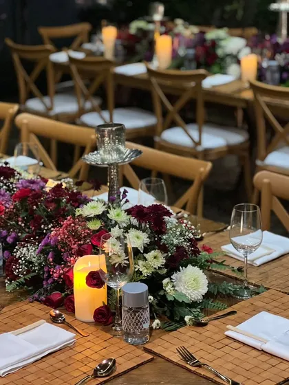A beautifully decorated sit-down dinner table. The combination of deep red and white flowers, candlelight, and rustic wooden chairs creates a warm and romantic ambiance.