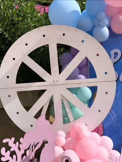 Details matter in creating a theme. This close-up shows a decorative ship's wheel prop against a custom "Pool Party" backdrop, surrounded by pink and purple balloons, adding to the nautical feel of the pre-wedding event.