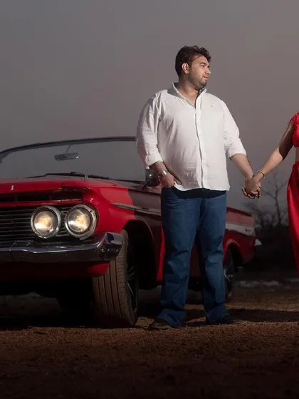 A beautiful evening shot of the couple holding hands in front of a vintage Chevrolet, with the car's headlights on.