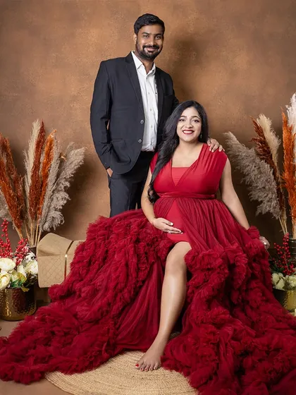 A full-length portrait of the couple in our rustic-themed setup. The mom-to-be is seated, looking radiant in a bold red gown, creating a powerful and romantic image.
