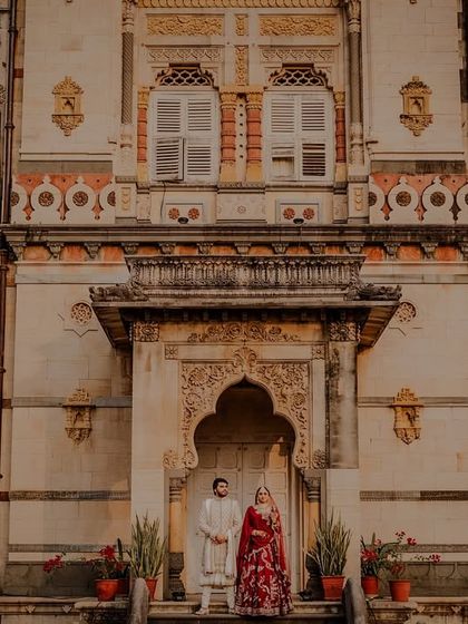 A wide shot showcasing the stunning facade of the palace with the couple standing in front.