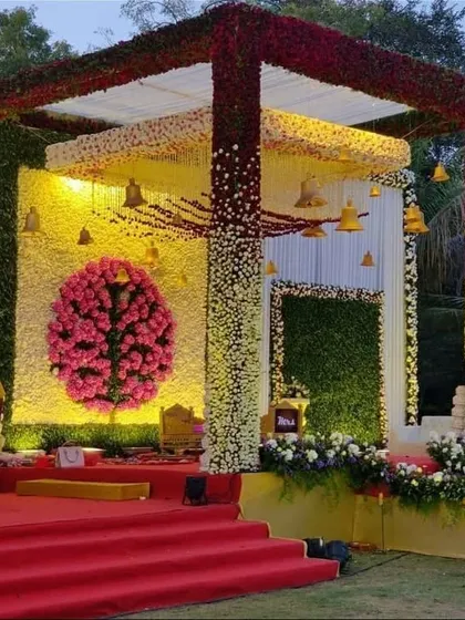 A vibrant and traditional floral mandap. The structure is covered in a gradient of red, yellow, and white flowers, with a Ganesha motif made of pink carnations as the centerpiece, creating a colorful and auspicious setting.