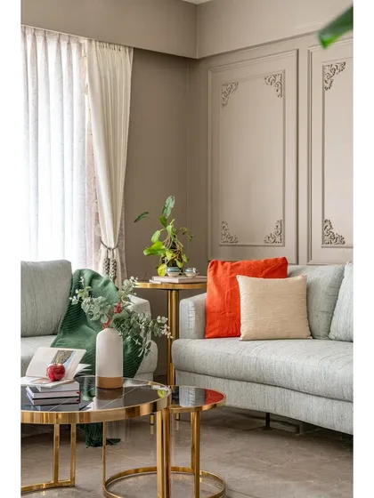 A close-up of the seating area, highlighting the mix of textures and colors. The vibrant orange and soft beige cushions add warmth to the cool grey sofa, complemented by the elegant gold-legged coffee table.