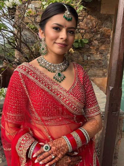 A radiant bride in a red lehenga with emerald jewelry. The makeup features a clean base, soft smokey brown eyes, and pink lips, creating a look that is both fresh and timeless for a destination wedding.