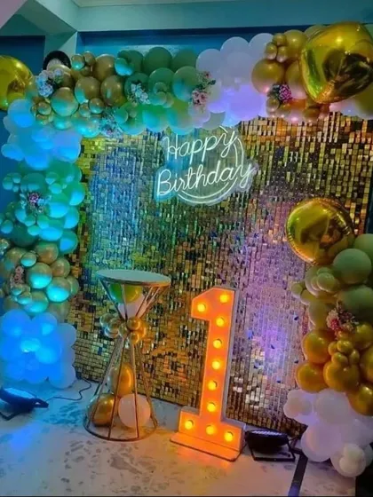 A glamorous first birthday setup with a gold sequin backdrop. The scene is framed by an organic balloon garland and features a "Happy Birthday" neon sign and a large light-up number "1."