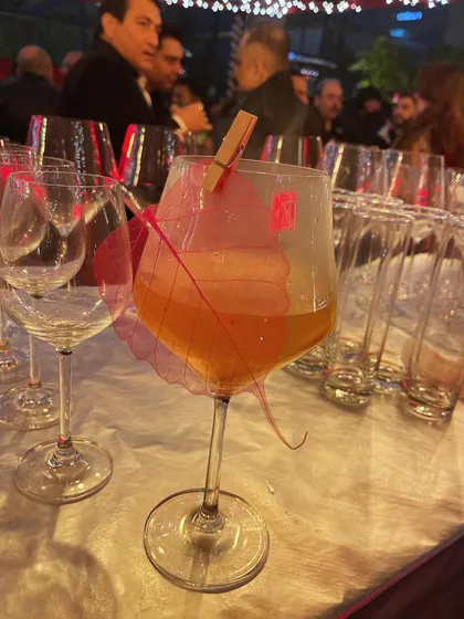 A sparkling cocktail served in a large wine glass, garnished with a translucent, colored leaf. The simplicity of the garnish allows the color and effervescence of the drink to shine.