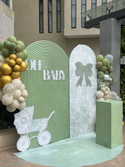 A modern and chic 'Oh Baby' shower setup in a lovely sage green. The decor features clean, arched backdrops with different textures and delicate line-art graphics of a bow and a baby carriage.