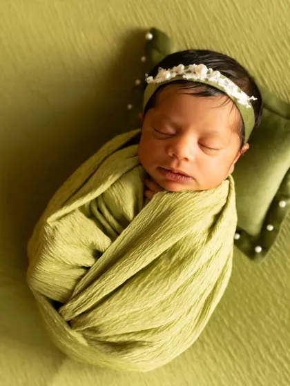 A close-up of a newborn swaddled in a textured olive green wrap, resting on a matching pillow with a delicate floral headband.