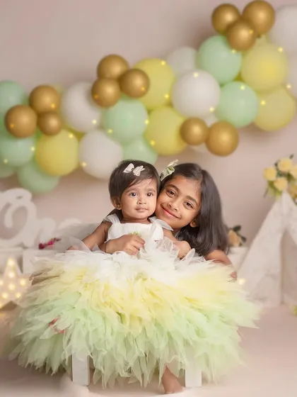 The sweet bond between sisters is a story I love to tell. Here, an older sister embraces her baby sibling during a milestone photoshoot, surrounded by soft pastel balloons and gentle light.