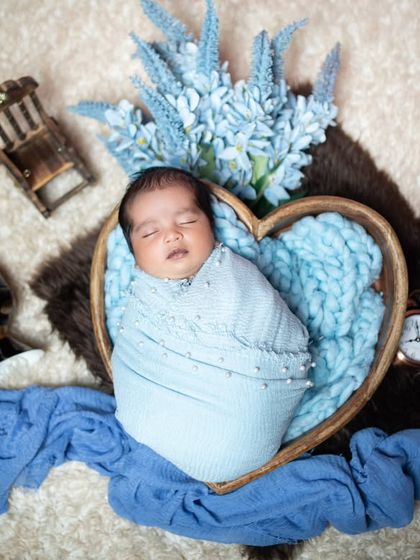 This newborn is sleeping in a heart-shaped basket, wrapped in a soft blue blanket. A sweet and serene portrait.