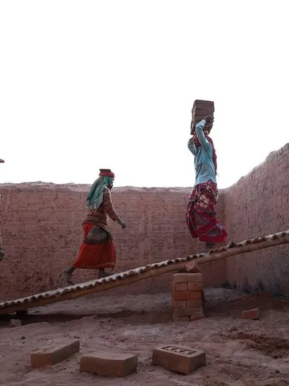 Women workers walk in a line across a makeshift bridge, each carrying a load of bricks. The image speaks to the communal nature of their labor.