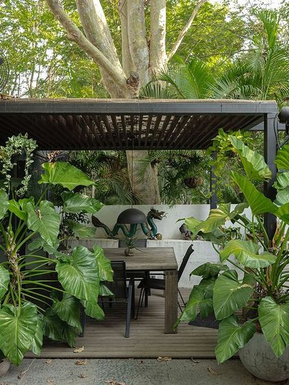 An outdoor dining area on a wooden deck, surrounded by oversized tropical plants. The space is designed for alfresco meals, with large planters creating a sense of enclosure and privacy.