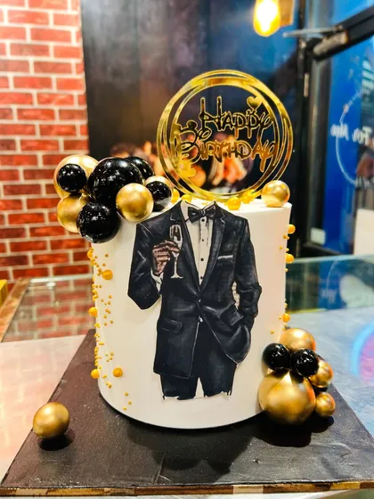A classy tuxedo-themed cake with an edible print, decorated with black and gold spheres for a sophisticated birthday.