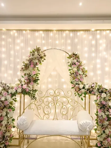 This is a classic and elegant seating arrangement for an engagement or roka. The white wrought-iron bench is framed by beautiful pink and white floral arrangements and a backdrop of warm fairy lights.
