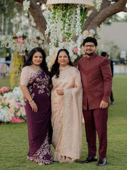 A family portrait from the reception. The siblings are styled in coordinating jewel tones, with the sister in a deep purple embroidered saree and the brother in a sharp maroon bandhgala.