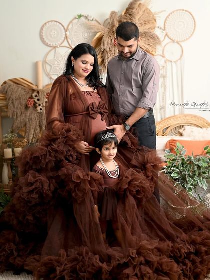 A gorgeous family maternity photo with a rich, earthy color palette. The mother and daughter wear matching ruffled brown gowns, creating a warm and stylish portrait against a boho-inspired set.