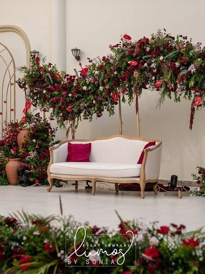 A regal reception stage designed with a stunning arch of deep red florals. The classic white sofa and terracotta pots add a touch of vintage elegance to the grand setting.