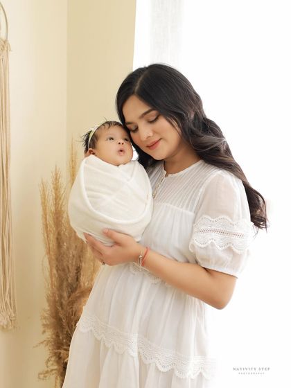 A mother gazes lovingly at her newborn, who is swaddled in pure white. The bright, airy setting with natural light creates a soft and angelic mood for this mother-child portrait.