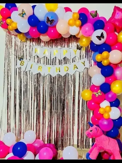 A simple and colorful birthday decoration for home. This setup includes a silver fringe backdrop, a "Happy Birthday" banner, and a multi-colored balloon arch.