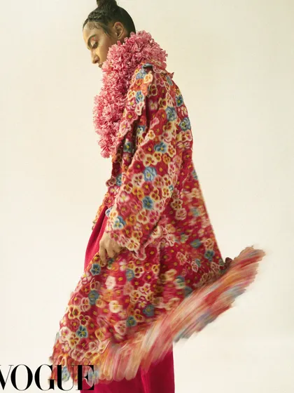 An editorial look for Vogue featuring a layered ensemble with a floral coat and a textured pink floral collar. The motion blur on the hem of the coat adds a dynamic quality, highlighting the rich textures and patterns of the designer pieces.