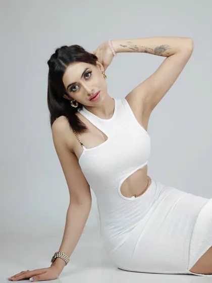 A full-body portrait of a model in a modern white dress, seated against a clean white background.