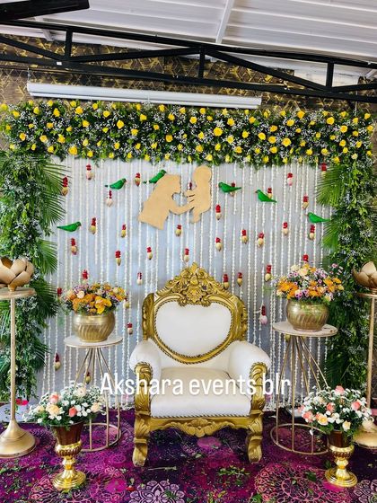 An elegant baby shower setup with a white backdrop featuring hanging jasmine and red flower buds. The decor is enhanced by parrot motifs and a central cutout symbolizing mother and child.